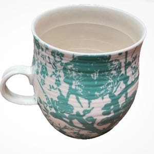 May include: A white ceramic mug with a rounded body and a curved handle. The mug features a teal floral pattern over a white background. The interior is white, and the mug has a capacity of approximately 12 ounces.