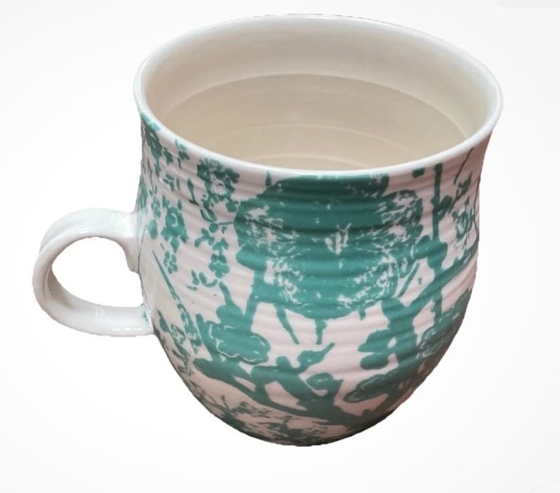 May include: A white ceramic mug with a rounded body and a curved handle. The mug features a teal floral pattern over a white background. The interior is white, and the mug has a capacity of approximately 12 ounces.