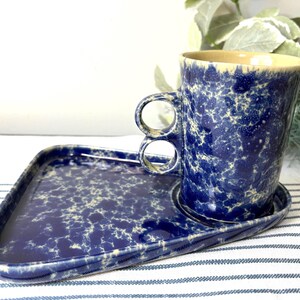 May include: A ceramic mug and matching triangular tray set featuring a deep blue and cream speckled glaze. The mug has a unique double-ring handle design, while the tray includes a circular indentation specifically shaped to hold the base of the mug.