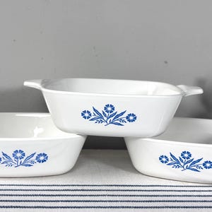 May include: Three white rectangular casserole dishes with handles, stacked. Each dish features a blue floral design. The dishes are on a striped surface, with a plant in the background. The dishes are likely for baking or serving.