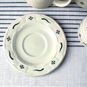 May include: A round, off-white ceramic saucer featuring a raised, segmented rim decorated with alternating dark green floral motifs and curved line accents. The center of the saucer is smooth and recessed, designed to hold a matching cup securely.