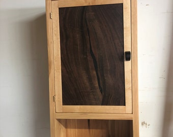 May include: A wooden wall-mounted cabinet with a single door and a shelf. The door is made of dark wood with a natural wood frame. The cabinet has a black knob.