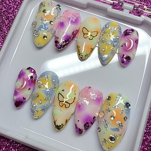 May include: A set of ten almond-shaped false nails in a white plastic case. The nails feature colorful designs with butterflies, moons, and sparkling embellishments. Colors include yellow, pink, purple, and silver. The background is a sparkly, magenta surface.