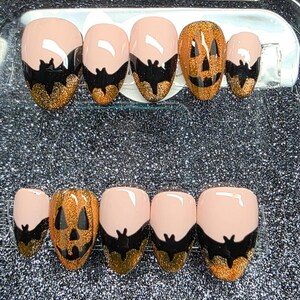 May include: A set of press-on nails in a clear plastic case. The nails feature a light pink base with black and gold accents. Some nails have a jack-o'-lantern design, while others have a bat design, perfect for Halloween.
