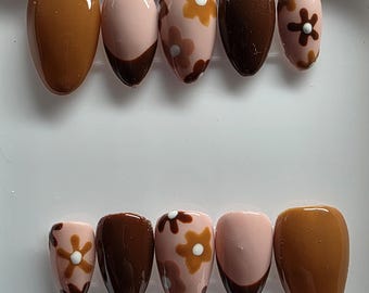 May include: A set of press-on nails in a white case. The nails feature a variety of designs, including solid brown, pink with brown tips, and pink with brown floral patterns. The nails are oval-shaped.