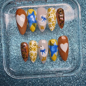 May include: A set of ten artificial nails in a clear plastic case. The nails feature various designs, including a white heart, a duck with a blue bow, and yellow floral patterns. One nail has the word "BREAD" written on it.