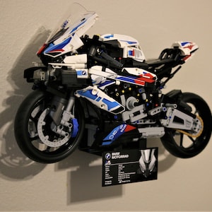 Könnte beinhalten: Ein weißes, blaues und rotes LEGO® BMW Motorrad Modellmotorrad mit einem schwarzen Ständer. Das Motorrad wird an einer weißen Wand ausgestellt. Das Motorrad hat eine schwarz-weiße Informationskarte mit dem Text "BMW Motorrad" darauf.