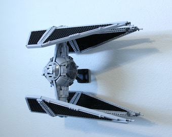 May include: A gray TIE fighter model with black wings, built with LEGO bricks. The model is mounted on a wall with a black stand.