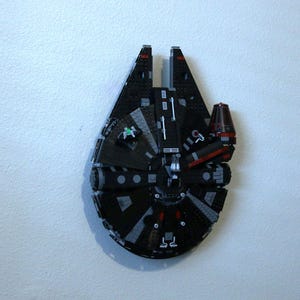 Puede incluir: Un modelo LEGO negro y gris del  Millennium Falcon. El modelo está construido en forma de la nave espacial icónica, con detalles como la cabina, los motores y el tren de aterrizaje. El modelo está montado en una pared blanca.