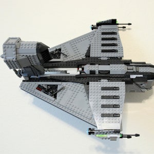 Puede incluir: Maqueta de nave espacial gris y negra construida con ladrillos de juguete entrelazados. El modelo presenta alas anguladas, m&uacute;ltiples torretas de ca&ntilde;&oacute;n y elementos estructurales detallados. El juguete est&aacute; sobre un fondo blanco.