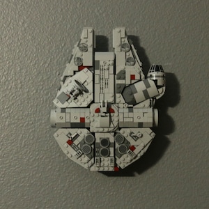 May include: A white and gray Lego Millennium Falcon spaceship model with red accents. The model is built with small plastic bricks and features a detailed design.