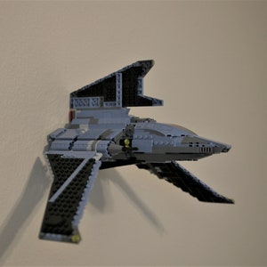 May include: A gray and black Lego spaceship with a detailed design. The spaceship has a pointed nose and two wings that are angled upwards. The spaceship is mounted on a wall.