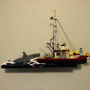 Puede incluir: Un modelo Lego de un barco de pesca con un tiburón atacándolo. El barco es rojo y blanco con un mástil amarillo y una pequeña tripulación de figuras Lego. El tiburón es gris y tiene la barriga blanca. El modelo está colocado sobre una base azul.
