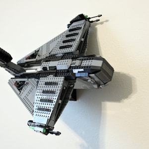 Puede incluir: Maqueta de nave espacial Lego gris y negra, montada en una pared blanca. El modelo presenta una construcci&oacute;n detallada con ladrillos Lego de varias formas y tama&ntilde;os. La nave espacial tiene m&uacute;ltiples alas y un cuerpo central, con peque&ntilde;os detalles verdes y negros.