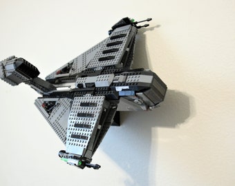 May include: A gray and black Lego spaceship model, mounted on a white wall. The model features detailed construction with various shapes and sizes of Lego bricks. The spaceship has multiple wings and a central body, with small green and black accents.