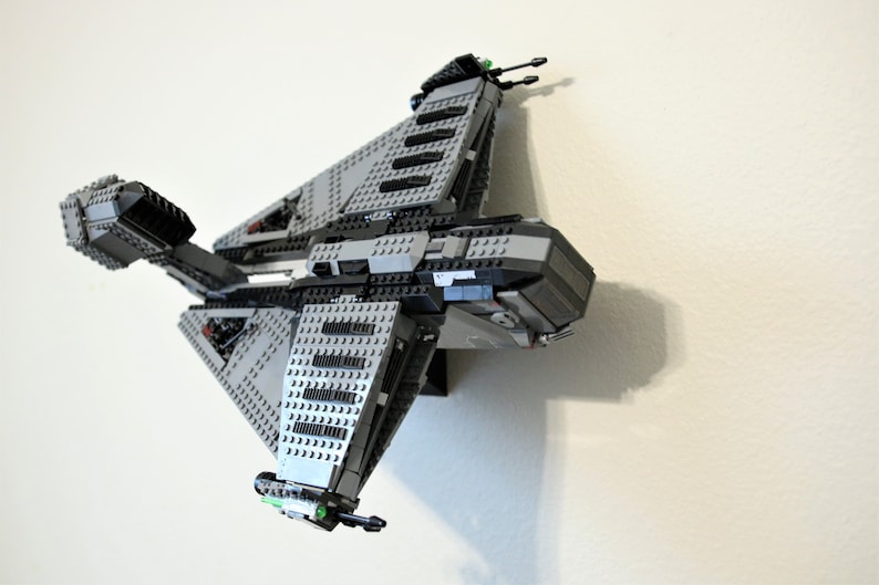 Puede incluir: Maqueta de nave espacial Lego gris y negra, montada en una pared blanca. El modelo presenta una construcci&oacute;n detallada con ladrillos Lego de varias formas y tama&ntilde;os. La nave espacial tiene m&uacute;ltiples alas y un cuerpo central, con peque&ntilde;os detalles verdes y negros.