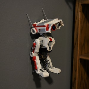 May include: A white and red Lego droid with a black body and red legs. The droid has a white head with a red stripe and two black eyes.