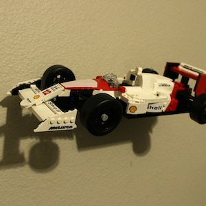 May include: A white and red Lego Formula 1 race car with black wheels and the words "Shell" and "McLaren" printed on the side. The car is suspended in mid-air, as if it is flying.