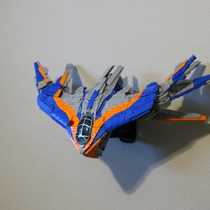 Puede incluir: Maqueta de nave espacial azul, naranja y gris, montada en una pared. El modelo presenta un diseño detallado con alas y una cabina. La nave espacial es un juguete o artículo de colección, probablemente de plástico.