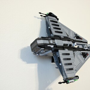 Puede incluir: Un modelo de nave espacial Lego gris y negro montado en una pared blanca. El modelo presenta una construcci&oacute;n detallada con varios ladrillos grises y negros, incluyendo alas anguladas y un cuerpo central. Se ven detalles verdes.