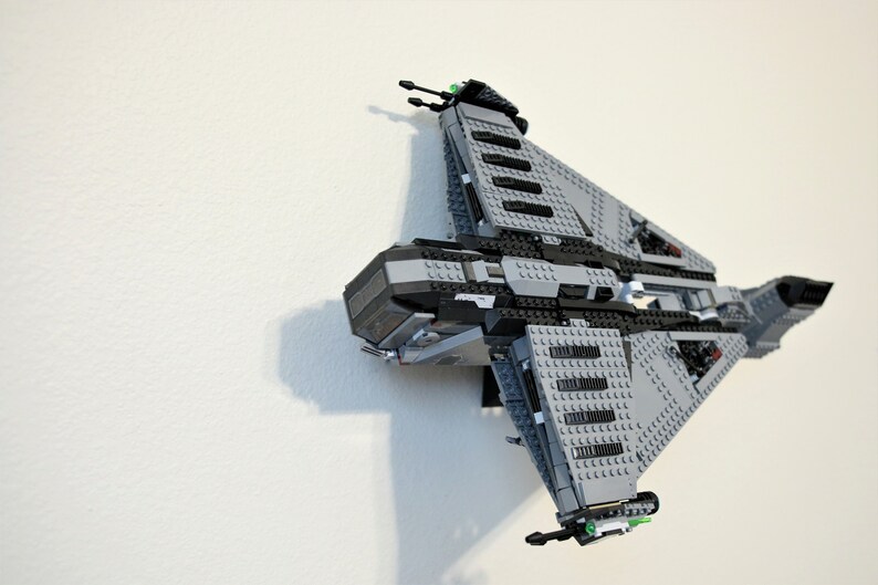 Puede incluir: Un modelo de nave espacial Lego gris y negro montado en una pared blanca. El modelo presenta una construcci&oacute;n detallada con varios ladrillos grises y negros, incluyendo alas anguladas y un cuerpo central. Se ven detalles verdes.