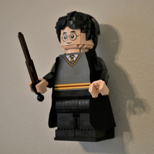 May include: A Lego minifigure of Harry Potter wearing a black robe and holding a brown wand. He has black hair, round glasses, and a grey sweater with a yellow tie.