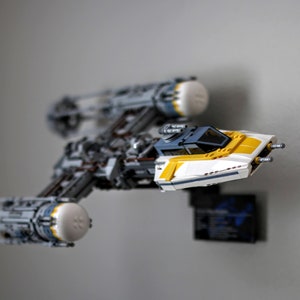 Peut inclure: Un chasseur X-wing Lego Star Wars blanc et jaune avec un cockpit bleu et des détails gris. Le chasseur est monté sur un mur.