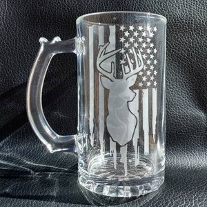 May include: Clear glass beer mug with a handle. The mug features a design of a deer silhouette superimposed over a stylized American flag. The flag design includes white stripes and a field of stars, creating a patriotic theme.