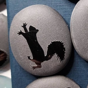May include: Gray, oval-shaped stone with a black silhouette of a squirrel in a playful pose. The squirrel is reaching upwards with its paws. The stone has a textured surface and is part of a collection.
