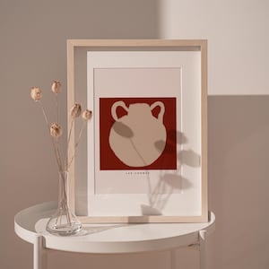 May include: Framed art print featuring a beige vase illustration on a dark red background. The print is in a light wood frame and includes the text "LES FORMES". The artwork is displayed on a white table.
