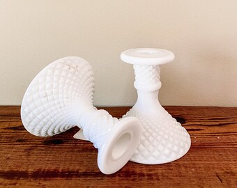 May include: Two white milk glass candle holders with a diamond pattern. The holders are on a wooden surface.