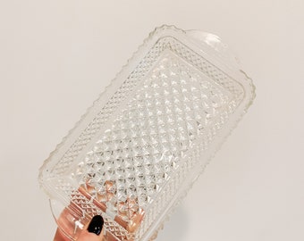 May include: A clear glass butter dish with a diamond pattern. The dish has two small handles on the sides.