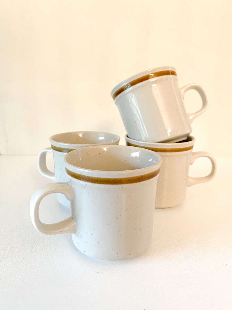 May include: Set of four white ceramic mugs with brown trim. The mugs have a speckled glaze and are stacked on top of each other.