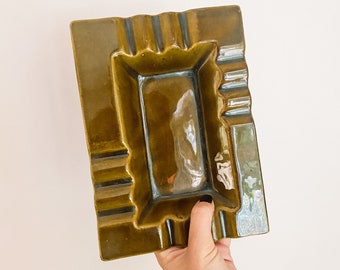 May include: A dark green ceramic ashtray with a rectangular shape and a raised rim. The ashtray has a smooth, glossy finish.