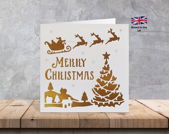 May include: A white Christmas card with a gold foiled design. The design features a sleigh with Santa Claus, reindeer, a Christmas tree, and the words "Merry Christmas".