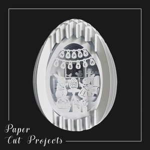 Op de afbeelding: Witte papieren uitsnijding van een paasei met een scène erin. De scène toont twee konijnen die een paasei versieren met een banner waarop "Happy Easter" staat.