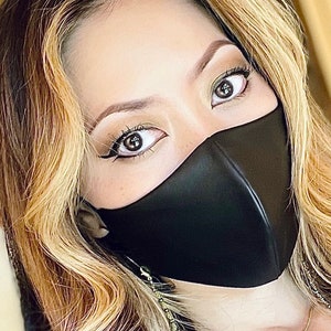 Puede incluir: Una mujer con el pelo largo y rubio lleva una mascarilla facial negra y un collar de oro de varias capas. El collar tiene un peque&ntilde;o colgante con la palabra "forever" en &eacute;l.