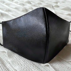 Puede incluir: Una mascarilla facial negra hecha de un tejido liso y satinado. La mascarilla tiene dos lazos para las orejas.