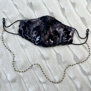 Puede incluir: Una mascarilla facial negra con un patr&oacute;n floral plateado y una cadena dorada con cuentas negras unidas a los lazos de las orejas.