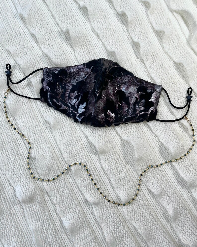 Puede incluir: Una mascarilla facial negra con un patr&oacute;n floral plateado y una cadena dorada con cuentas negras unidas a los lazos de las orejas.