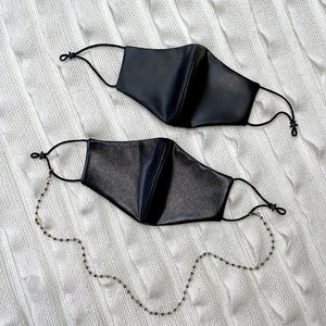 Puede incluir: Dos mascarillas negras con una cadena dorada. La mascarilla de arriba est&aacute; etiquetada como "Black Vegan Leather" y la de abajo como "Black Shiny Vegan Leather".