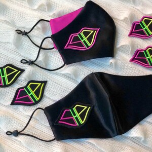 May include: Two black satin face masks with pink and green lips embroidered on them. The masks have black elastic ear loops. There are three extra embroidered lips on a white surface.