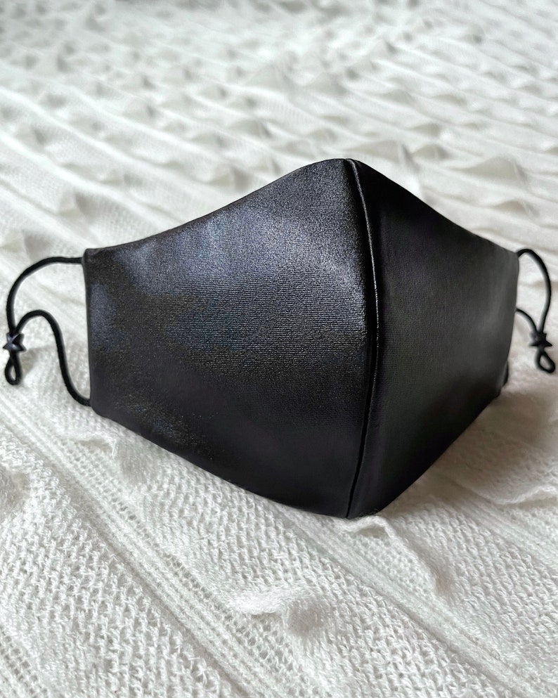 Puede incluir: Una mascarilla facial negra hecha de un tejido liso y satinado. La mascarilla tiene dos lazos para las orejas.