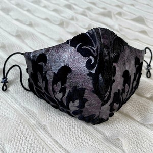 Puede incluir: Una mascarilla facial negra y plateada con un patr&oacute;n de damasco. La mascarilla tiene correas el&aacute;sticas negras.