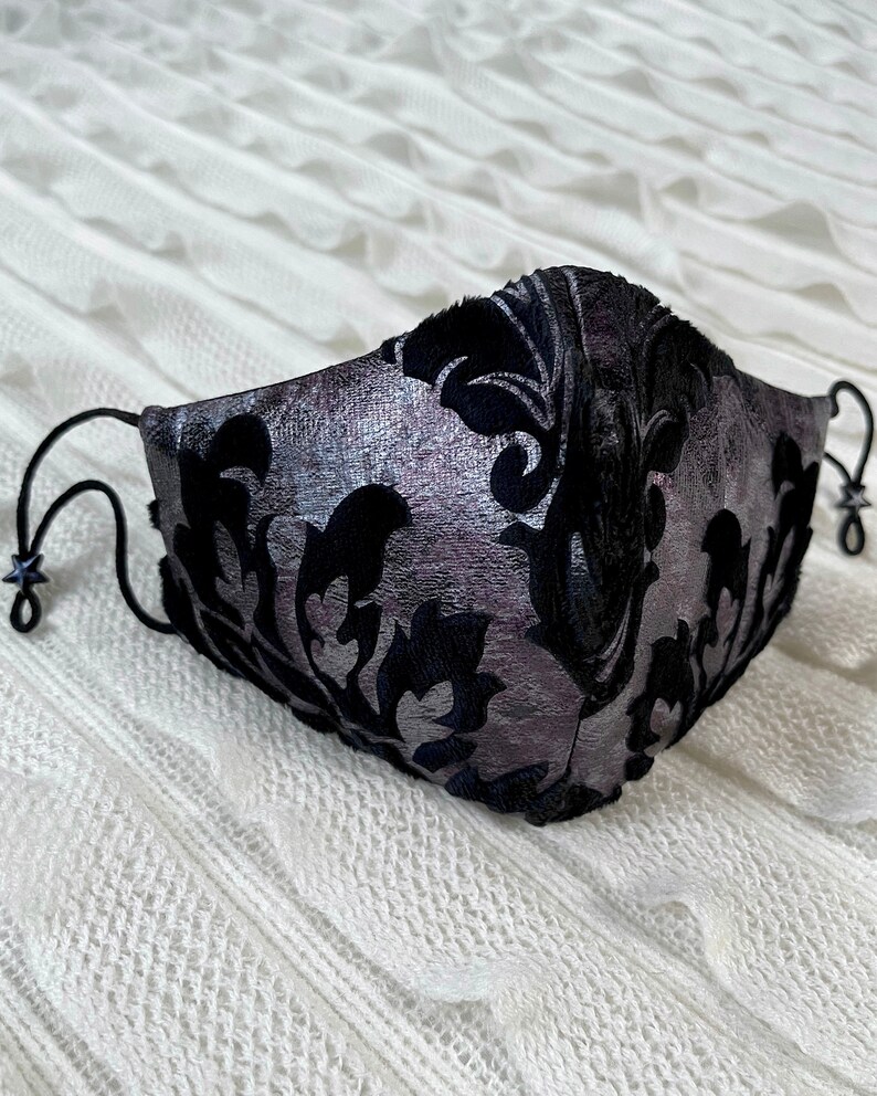 Puede incluir: Una mascarilla facial negra y plateada con un patr&oacute;n de damasco. La mascarilla tiene correas el&aacute;sticas negras.