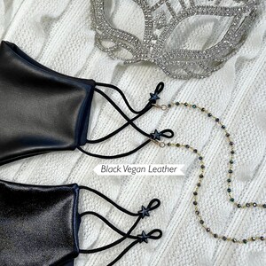Puede incluir: Dos mascarillas negras con cierres en forma de estrella negra y correas el&aacute;sticas negras. La mascarilla superior est&aacute; etiquetada como "Black Vegan Leather" y la inferior como "Black Shiny Vegan Leather". Se muestra una cadena dorada con cuentas negras junto a las mascarillas.