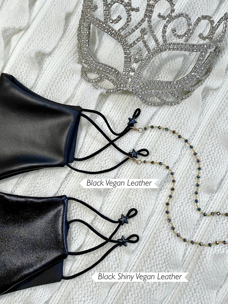 Puede incluir: Dos mascarillas negras con cierres en forma de estrella negra y correas el&aacute;sticas negras. La mascarilla superior est&aacute; etiquetada como "Black Vegan Leather" y la inferior como "Black Shiny Vegan Leather". Se muestra una cadena dorada con cuentas negras junto a las mascarillas.
