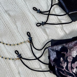 Puede incluir: Dos mascarillas negras con topes de cord&oacute;n en forma de estrella negra y una cadena dorada con cuentas negras. Las mascarillas est&aacute;n sobre una superficie blanca y peluda.