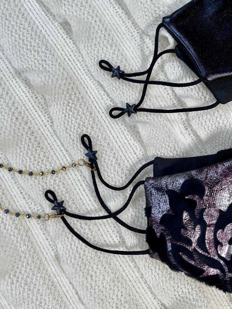 Puede incluir: Dos mascarillas negras con topes de cord&oacute;n en forma de estrella negra y una cadena dorada con cuentas negras. Las mascarillas est&aacute;n sobre una superficie blanca y peluda.