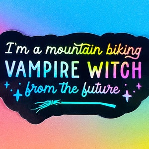 May include: Black sticker with the text "I'm a mountain biking VAMPIRE WITCH from the future" in a rainbow gradient. The sticker has a witch's broom and star accents. The background is a gradient of blue, pink, and yellow.